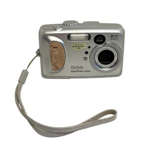 以下が含まれることがあります： 2.0メガピクセルのシルバーKodak EasyShare CX6230デジタルカメラ。カメラにはレンズ、フラッシュ、リストストラップが付いています。「Kodak Color Science Inside」と「Kodak EasyShare CX6230」の文字が見えます。