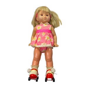 May include: A doll with blonde hair and a pink dress featuring a white daisy pattern and a bright pink waistband. The doll wears white socks with pom-poms and red roller skates with black wheels. The doll has a light skin tone and a pink headband.