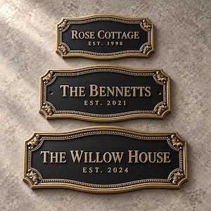 Puede incluir: Tres placas de pared rectangulares con bordes dorados ornamentados y centros negros. La placa superior dice ROSE COTTAGE EST. 1998, la del medio THE BENNETTS EST. 2021 y la inferior THE WILLOW HOUSE EST. 2024 en letras doradas en relieve.