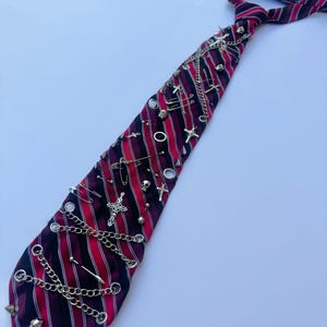 May include: A necktie featuring a diagonal striped pattern in red, black, and white. The tie is embellished with various silver-toned metal hardware, including small skull charms, cross pendants, safety pins, circular rings, and draped link chains.