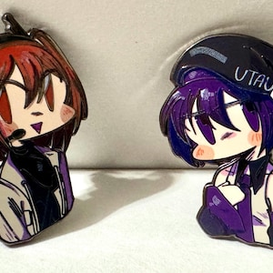 May include: Two enamel pins featuring anime-style characters. The left pin shows a character with red hair and a headset. The right pin shows a character with purple hair, a dark cap with the text UTAU, and blue headphones with white accents.