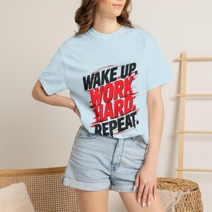 May include: Light blue t-shirt with the text "WAKE UP. WORK HARD. REPEAT." in black and red. The shirt is paired with denim shorts. The overall theme is fitness and motivation.