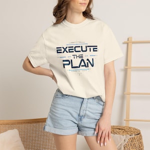 May include: Cream-colored t-shirt with the text "EXECUTE THE PLAN" in blue and white. The shirt is a crew neck and is paired with denim shorts. The design features a modern, tech-inspired font.