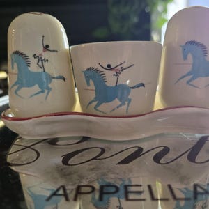 May include: A ceramic condiment set featuring two rounded shakers and a central open container on an elongated tray. Each piece is cream-colored with a blue graphic of a horse and a figure performing a circus-style stunt on its back.