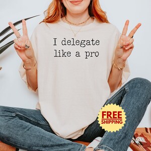 May include: A cream-colored t-shirt with the text "I delegate like a pro" in black. The person is wearing blue jeans and making a peace sign with both hands. A yellow sticker with the words "FREE SHIPPING" is visible.