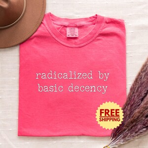 以下が含まれることがあります： 「radicalized by basic decency」の白文字がプリントされた、コーラル色のTシャツ。クルーネックで、中央上部にタグが付いています。茶色の帽子とドライフラワーが背景にあります。