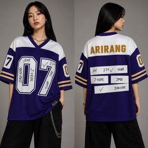 Op de afbeelding: Een paars en wit shirt in jersey-stijl met het nummer "07" op de voor- en achterkant. De voorkant heeft de namen "JUNG KOOK", "J-HOPE" en "JIN". De achterkant zegt "ARIRANG" en vermeldt namen als "SUGA" en "JIMIN".