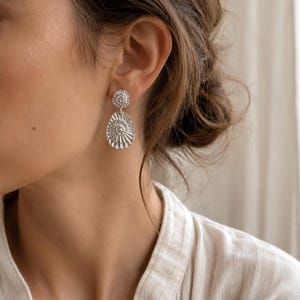 May include: A pair of silver-toned drop earrings featuring a small circular stud connected to a larger, oval-shaped pendant. Both components display a textured, radiating spiral pattern reminiscent of an ammonite fossil shell design.