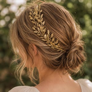 Puede incluir: Un peine para el cabello de color dorado con forma de rama con hojas, usado en el cabello. El peine es de metal y tiene detalles intrincados. El cabello está peinado en un moño bajo. El fondo está borroso con follaje verde.