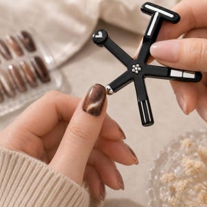 Könnte beinhalten: Ein schwarzes Nagelkunstwerkzeug mit einem T-förmigen Griff und einem sternförmigen Design. Das Werkzeug wird verwendet, um einen braunen und weißen Nagel aufzutragen. Ein Set brauner Nägel ist im Hintergrund sichtbar.