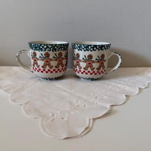 May include: Two ceramic mugs with a gingerbread man design. The mugs have a white base with a red and white checkered pattern at the bottom. The top rim is green with white dots. The mugs are on a white, scalloped-edge cloth.
