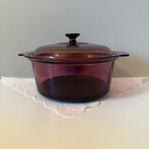May include: A deep, round, translucent plum-colored glass casserole dish with integrated side handles and a matching glass lid featuring a central knob handle. The vessel has a smooth, polished finish and a wide, open circular opening at the top.