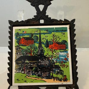 May include: Black metal trivet with a detailed landscape tile. The tile features a church, houses, and a waterfall set against a green and blue background. An eagle tops the trivet.