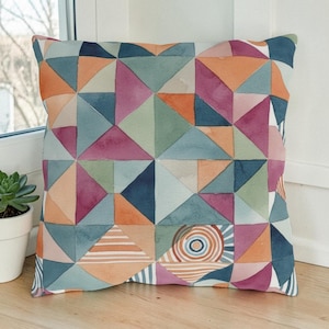 May include: Square decorative pillow with a geometric watercolor design. The pillow features a patchwork of triangles in shades of blue, orange, pink, and green. A circular design with concentric rings is visible on the lower right corner.