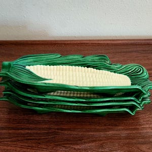 May include: A stack of four ceramic corn-shaped serving dishes. Each dish features a central pale yellow section with a textured kernel pattern, surrounded by deep green, molded ceramic husks that form the edges and handles of the tray.