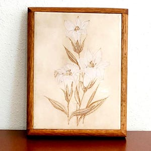 May include: A framed floral artwork featuring white lilies with brown stems and leaves. The lilies are painted on a cream-colored background, and the artwork is encased in a wooden frame.