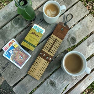 Op de afbeelding: Een bruin leren cribbagebord met metalen details, een pakje "Star" gouden speelkaarten en twee kopjes koffie. De items zijn geplaatst op een verweerd houten oppervlak, wat een informele setting suggereert.