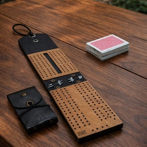 Op de afbeelding: Een houten cribbagebord met een zwarte leren bovenkant en een kleine zwarte leren kaarthouder. Een pak speelkaarten ligt naast het bord. Het woord "START" staat op het bord.