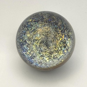 May include: A spherical object with a smooth, clear surface containing a dense, swirling interior pattern. The core features a mix of metallic gold, deep blue, and silver flecks that create a textured, galaxy-like appearance within the sphere.