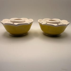 May include: Two yellow ceramic flower-shaped bowls with a white rim and center. The bowls have a circular opening in the center and small holes around the edge. The bowls are on a white surface.