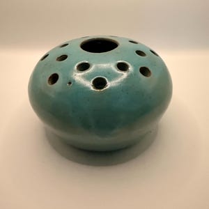 May include: A teal-colored ceramic vase with a rounded shape and multiple circular openings. The vase has a glossy finish and a dark interior. The openings are evenly spaced around the top, suggesting a design for floral arrangements.