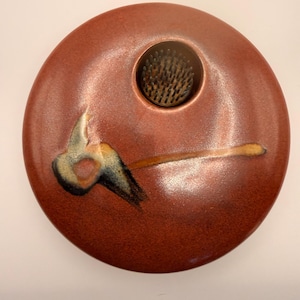 May include: A round, reddish-brown ceramic flower frog with a central hole containing a kenzan. The frog has a painted design with black, yellow, and blue accents. The overall design is a minimalist, artistic style.