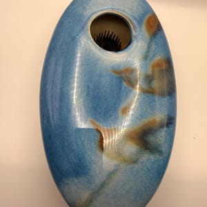 May include: Oval-shaped ceramic vase in shades of blue with brown and white accents. The vase has a circular opening at the top and a textured interior. The vase is designed for floral arrangements.