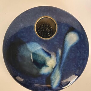 May include: A circular ceramic flower frog with a glossy deep blue glaze featuring mottled black and white accents. The piece has a central circular opening containing a metal pin holder with numerous upright needles for arranging flowers.