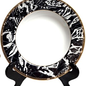 May include: A round ceramic plate featuring a solid white center and a wide border decorated with a black and white marble pattern. The outer edge of the plate is finished with a thin gold rim and a delicate gold beaded detail along the inner border.