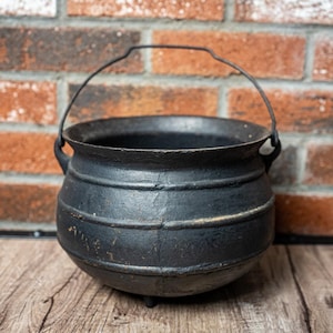 May include: A black cast iron cauldron with a rounded body, three small feet, and a thin metal wire handle attached to two side lugs. The exterior surface features horizontal ridges and a textured finish, showing signs of wear and age.