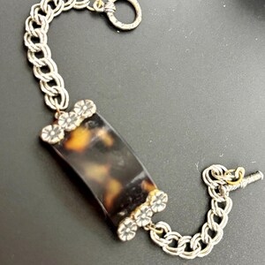 May include: A bracelet featuring a rectangular, mottled brown and black tortoiseshell-patterned center piece flanked by two silver-toned metal floral accents. The piece is connected to a silver-toned chain with a toggle clasp closure.