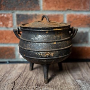 May include: A black cast iron pot with three legs and a matching lid. The pot features a rounded body with horizontal ridges and a small handle on each side. The word "MALON" is embossed on the front of the pot, which shows signs of wear.