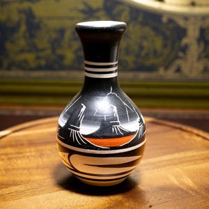 Può includere: Un piccolo vaso in ceramica bulboso con finitura nera opaca, caratterizzato da motivi geometrici dipinti a mano in bianco e arancione e motivi stilizzati di uccelli. Il collo è decorato con strisce bianche orizzontali e la base è arrotondata.