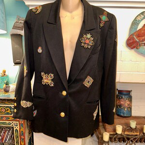 May include: Black blazer adorned with colorful jeweled embellishments. The blazer features gold buttons and decorative patches with intricate designs. A classic tailored jacket with a unique, artistic flair.