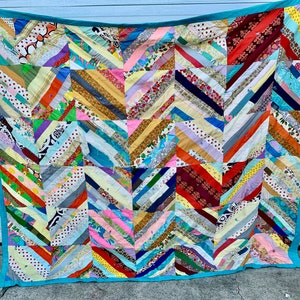May include: A rectangular quilt featuring a patchwork design of multi-colored fabric strips arranged in diagonal patterns. The quilt is bordered by a solid turquoise fabric trim and consists of various floral, polka dot, and geometric prints.