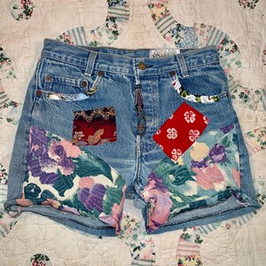May include: Blue denim shorts featuring a high-waisted cut, a button fly, and various fabric patches. The design includes floral-patterned panels on the lower legs, a red square patch with white flowers, and a dark patterned rectangular patch.