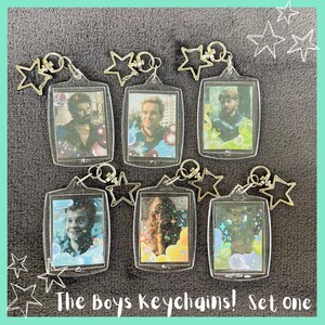 Puede incluir: Un conjunto de seis llaveros rectangulares de acrílico transparente con retratos de personajes de The Boys, cada uno unido a un cierre plateado en forma de estrella. Los llaveros incluyen superposiciones holográficas de estrellas y burbujas, con el texto The Boys Keychains! Set one.