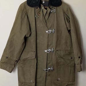 May include: Olive green coat with a black collar. The coat features large pockets, metal clasps, and a button closure. The coat is hanging on a wooden hanger against a neutral background.