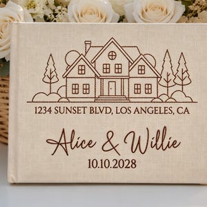 Könnte beinhalten: Ein rechteckiges, cremefarbenes, stoffbezogenes Fotoalbum mit einer dunkelbraunen, gestickten Strichzeichnung eines Hauses mit Bäumen. Der Text lautet 1234 SUNSET BLVD, LOS ANGELES, CA, Alice & Willie und das Datum 10.10.2028.