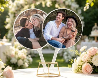 Puede incluir: Dos abanicos fotográficos circulares con imágenes de una pareja, cada uno unido a un mango de plástico transparente. Los abanicos se muestran en posición vertical en un soporte de alambre dorado. El abanico izquierdo muestra a una pareja sonriendo.