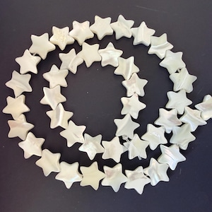 Può includere: Un filo di perline bianche a forma di stella con una lucentezza perlescente. Le perline sono disposte in una linea curva su uno sfondo scuro. Le stelle hanno cinque punte e sono di dimensioni uniformi.