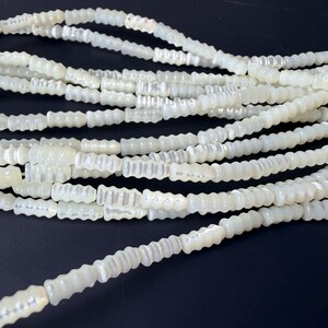 Peut inclure: Gros plan sur plusieurs brins de perles nacrées blanc cassé. Les perles sont cylindriques avec un motif cannelé. Elles sont enfilées, idéales pour la fabrication de bijoux ou l'artisanat. Les perles sont présentées sur un fond sombre.