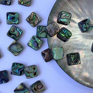 May include: Square abalone shell tiles in shades of green, blue, and brown are scattered on a white surface and near a mother-of-pearl shell. The tiles have a polished, reflective surface, suitable for jewellery making or craft projects.