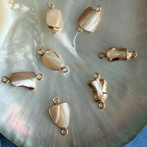 May include: Seven gold-toned jewelry connectors with iridescent, off-white inlays. The connectors have a variety of shapes, including rectangular, oval, and irregular, with small loops at each end for attaching to other jewelry components.