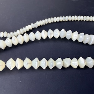 May include: Three strands of white, iridescent beads of varying sizes against a dark background. The beads have a pearlescent sheen, with some shaped like elongated diamonds and others round.