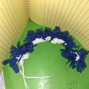 Puede incluir: Una pieza de ganchillo hecha a mano con forma curva, que utiliza hilo azul y blanco. El hilo azul crea un efecto texturizado y con flecos, mientras que el hilo blanco forma una base contrastante. La pieza se muestra sobre una superficie verde.