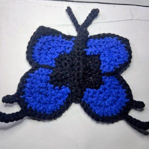 May include: A handcrafted crochet butterfly in blue and black. The butterfly has detailed wings and antennae, displaying a textured, handmade design. This decorative item is suitable for various craft projects.