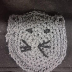 May include: A gray crocheted cat face patch featuring a circular shape with two small pointed ears at the top. The facial features, including two horizontal eyes and a set of whiskers, are embroidered in black yarn onto the gray crocheted surface.