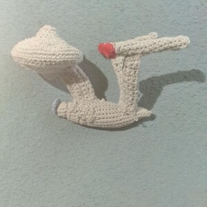 May include: A handmade, off-white crochet Star Trek Enterprise model. The crochet spaceship features a mushroom-shaped head, two extended wings, and a small red heart detail. The craft is displayed against a light blue wall.