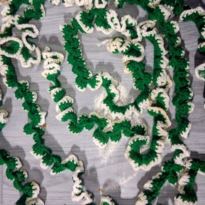 May include: A long, spiraling crochet garland featuring a repeating pattern of deep forest green yarn centers bordered by a ruffled, cream-colored edge. The decorative textile strand is coiled in a loose, circular arrangement with a wavy texture.